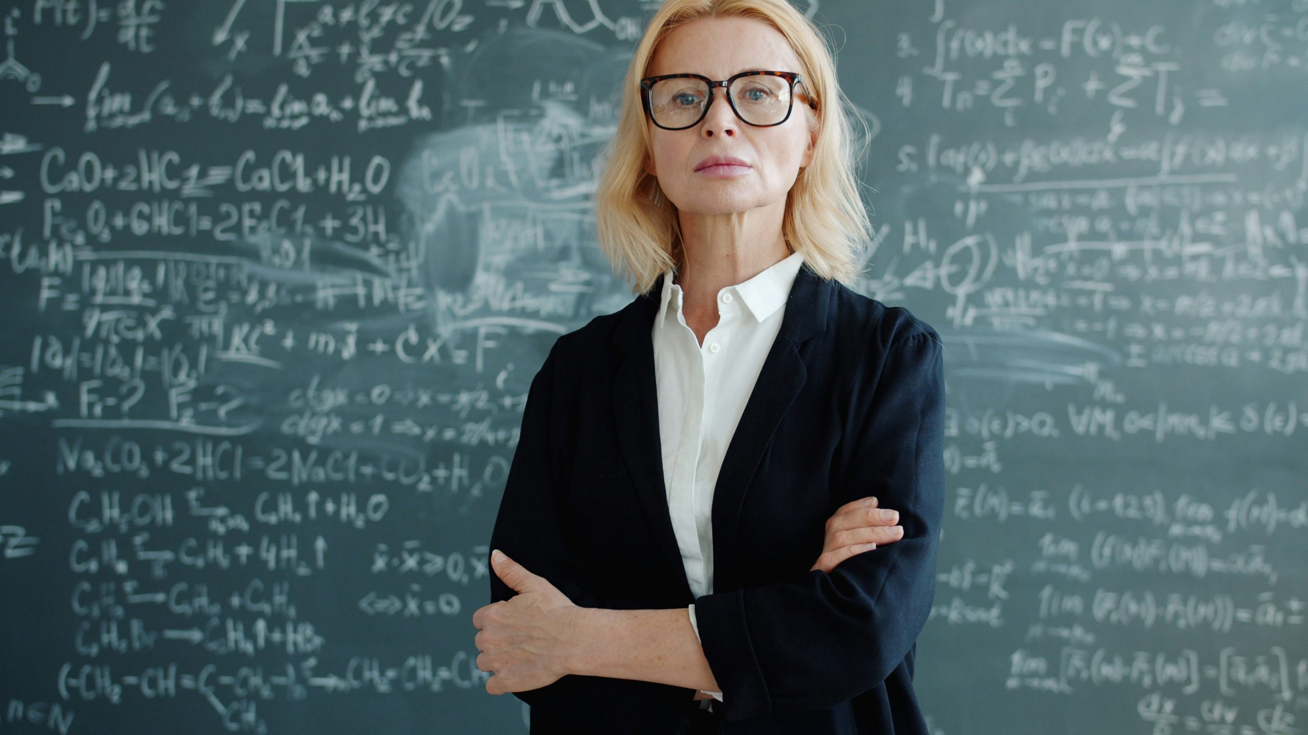 Portrait of serious smart lady professor in classroom with formulas on chalkboard standing alone with arms crossed looking at camera. People and education concept.